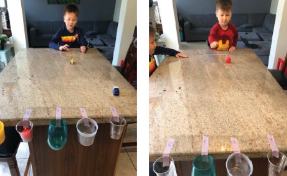 Two boys sit at a table and roll objects into plastic cups taped to the end of the table