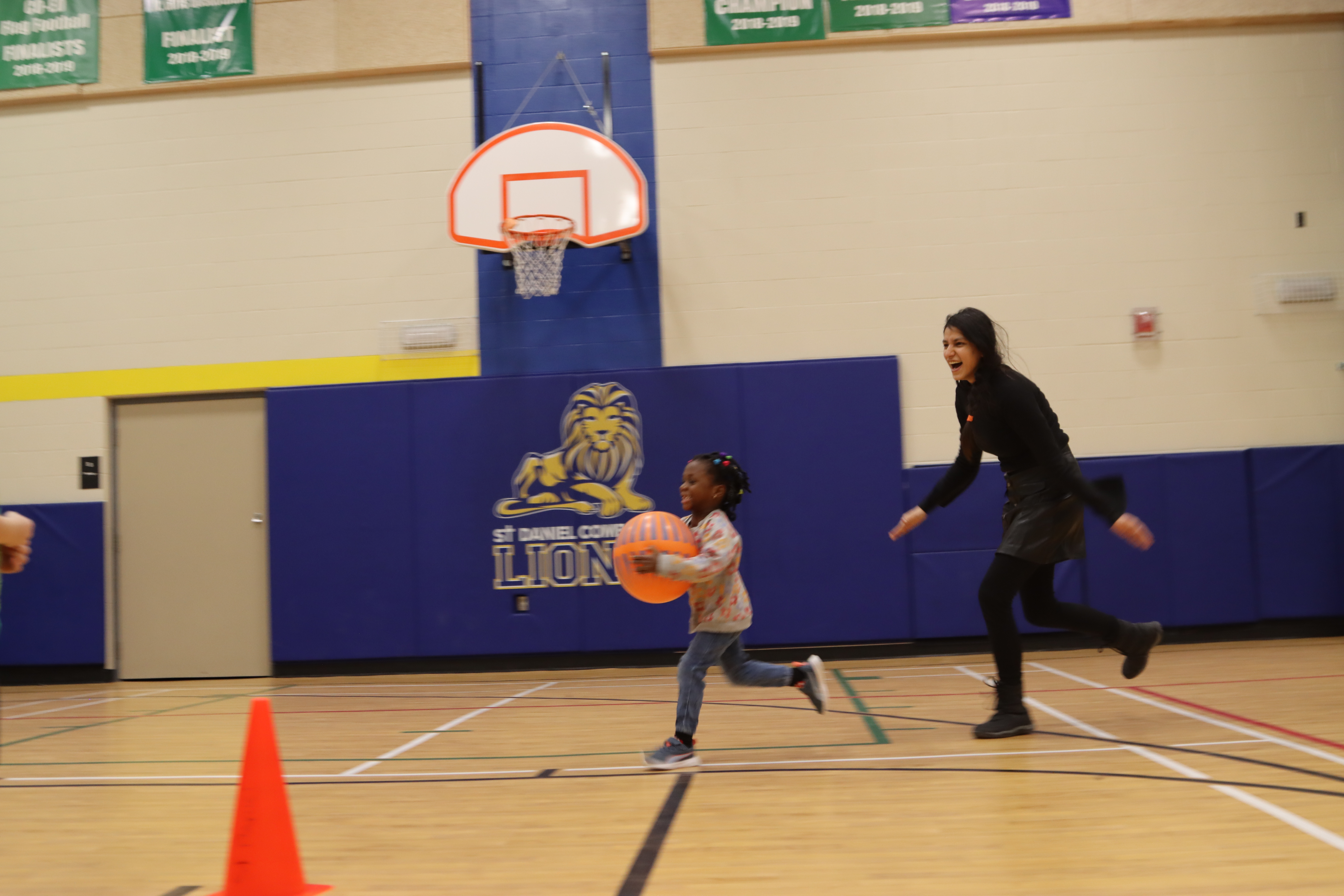 A young girl and educator in a PLASP after school program runs with an oversize basketball in a school gym