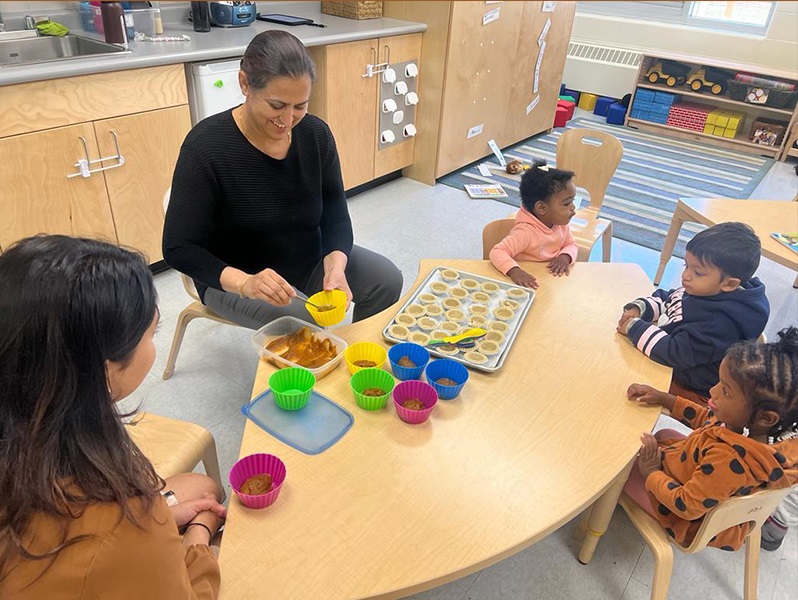 Two PLASP educators sit at a table in a program classroom and prepare snacks for three toddlers