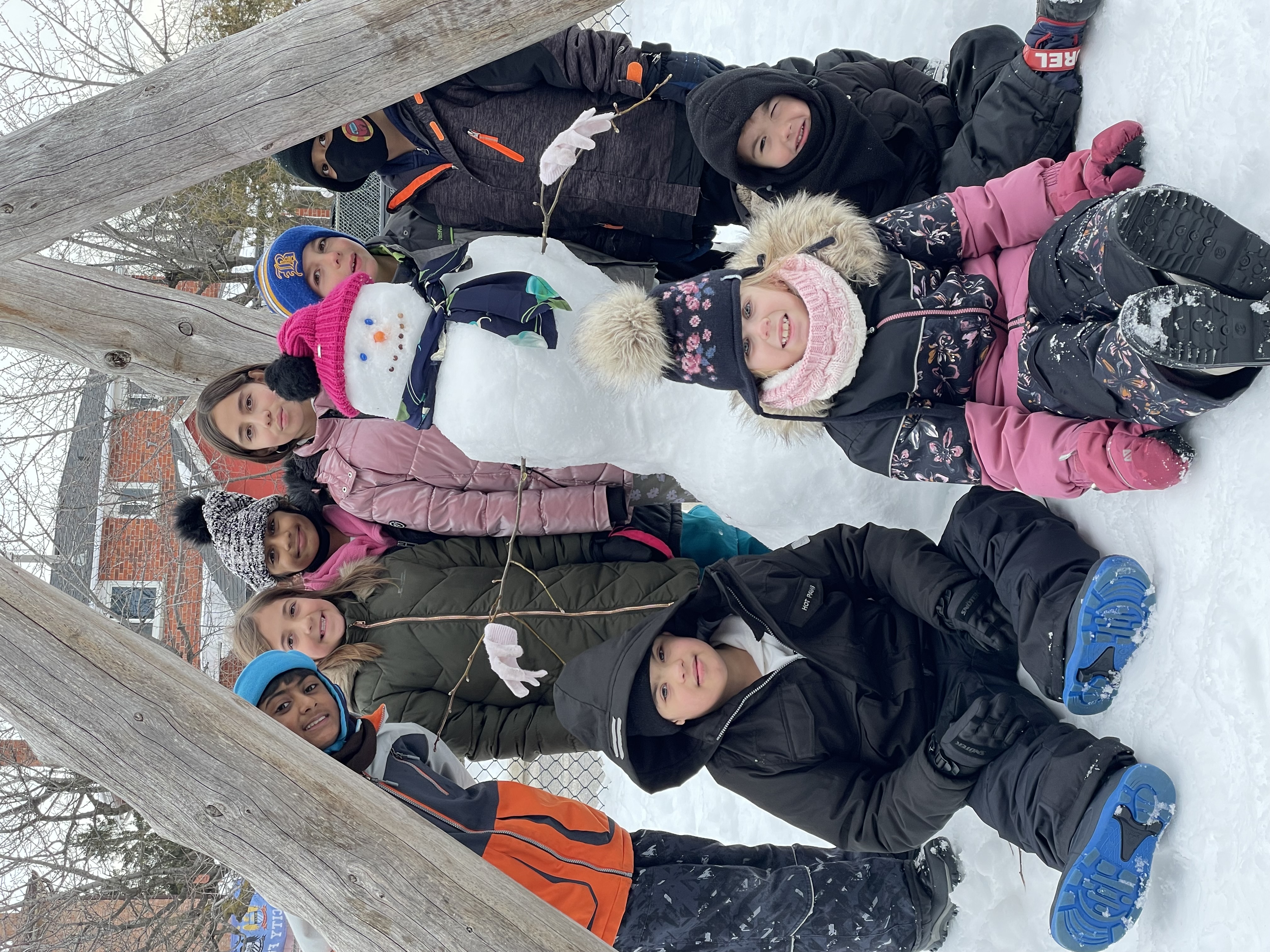A group of children dressed in snow gear stand around the snowman they built at a PLASP program