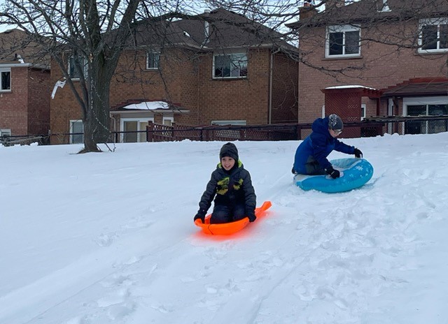 Two boys sled down a snow-covered hill, one on a sled and one in a tube
