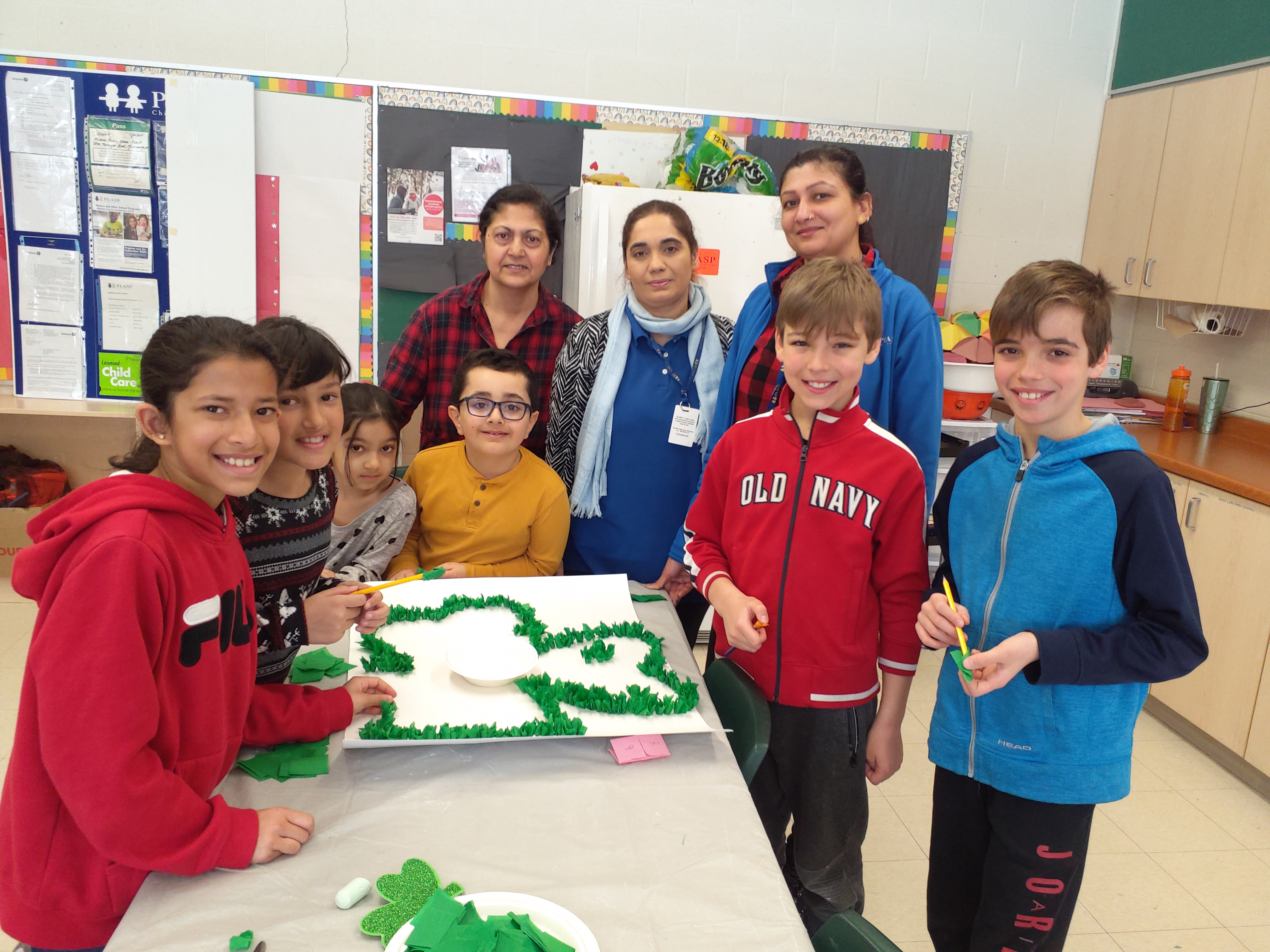 Three PLASP educators stand around a table with children in their program, who are proudly displaying their artwork